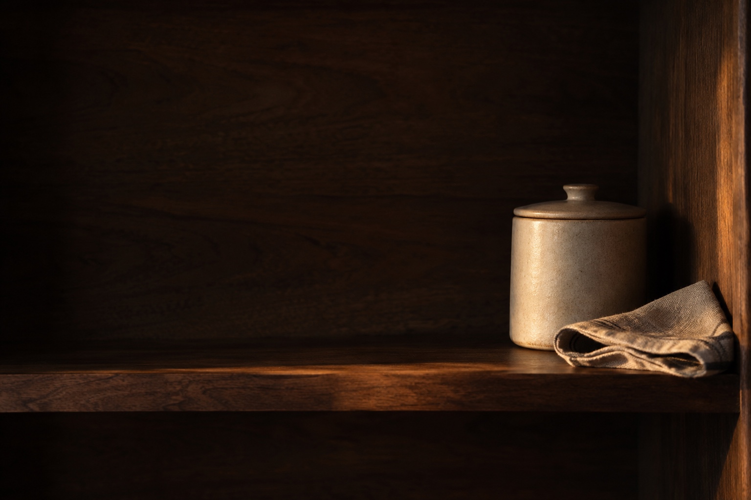 A ceramic jar and folded linen on a walnut pantry shelf in the last warm light of day